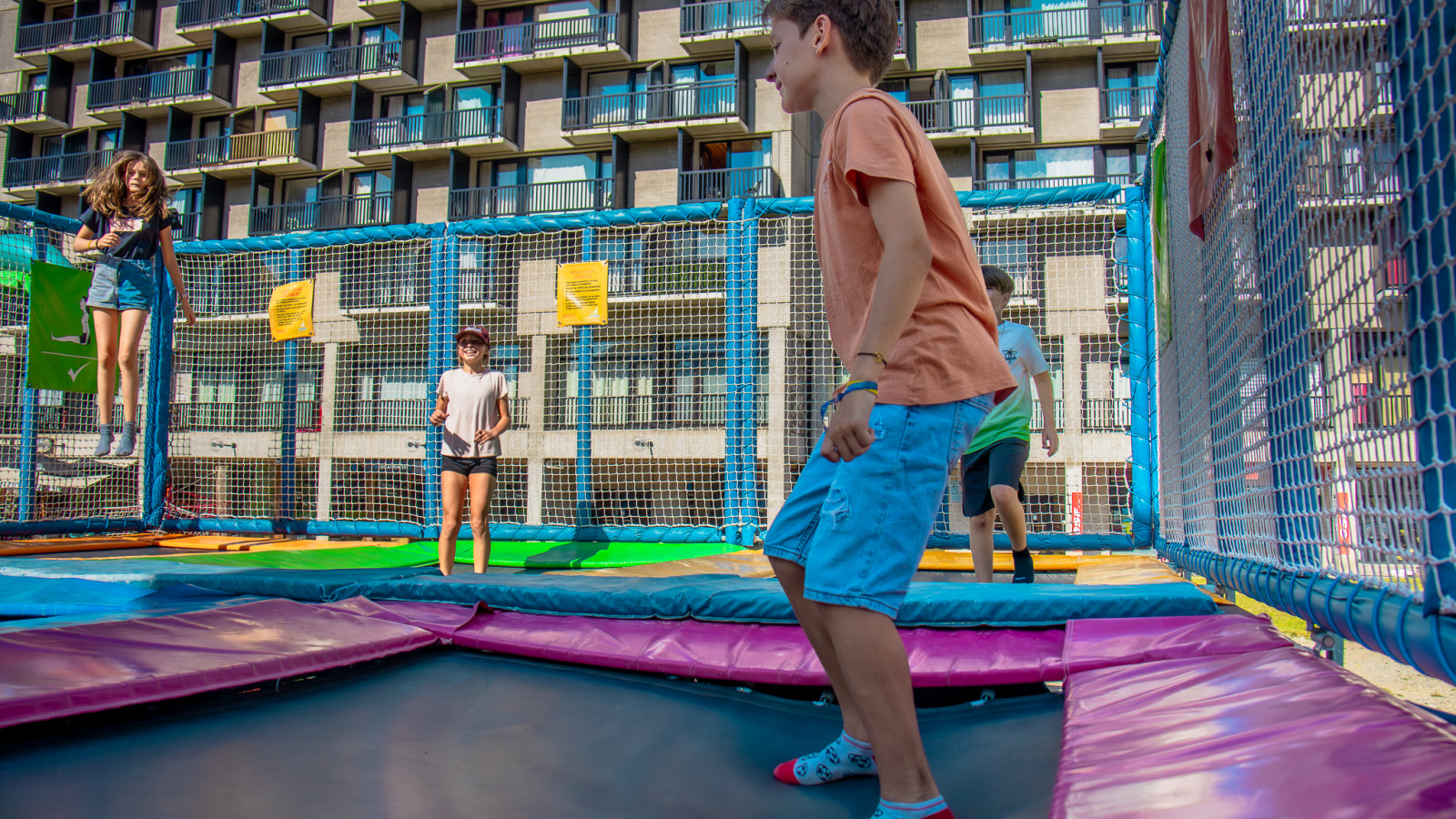 Several children each playing on one of the six trampoline mats, with one of the buildings in the Flaine resort in the background