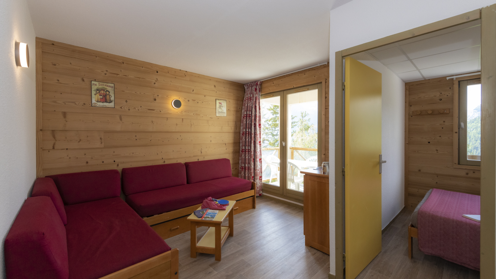 Appartement avec un grand canapé, salon donnant sur le balcon où se trouve une table de jardin et des chaises. Documentation sur la table de salon. Chambre donnant sur une vue montagne avec un lit double.