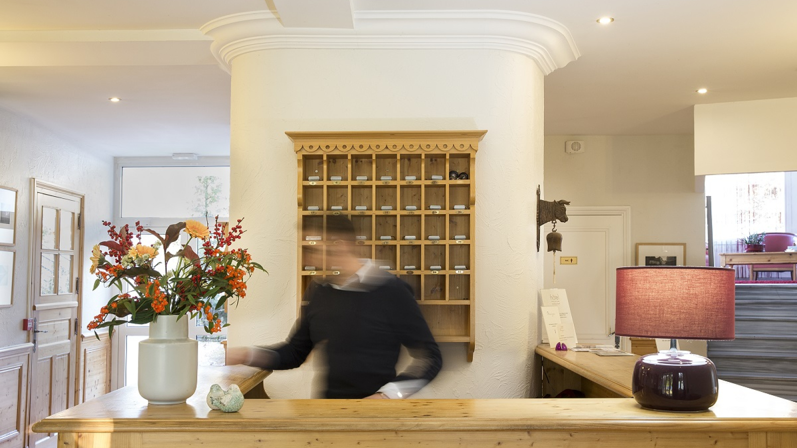 Réception d'hôtel lumineuse et accueillante. Derrière un grand comptoir en bois clair, on peut apercevoir une personne souriante. Des fleurs colorées dans un vase apportent une touche de fraîcheur à l'espace.