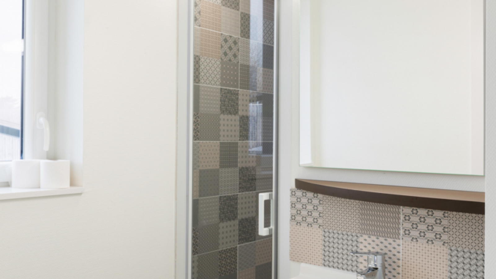 Une salle d'eau présente un lavabo immaculé sous un miroir, à côté d'une cabine de douche. On devine la présence d'une fenêtre auprès de la cabine de douche.