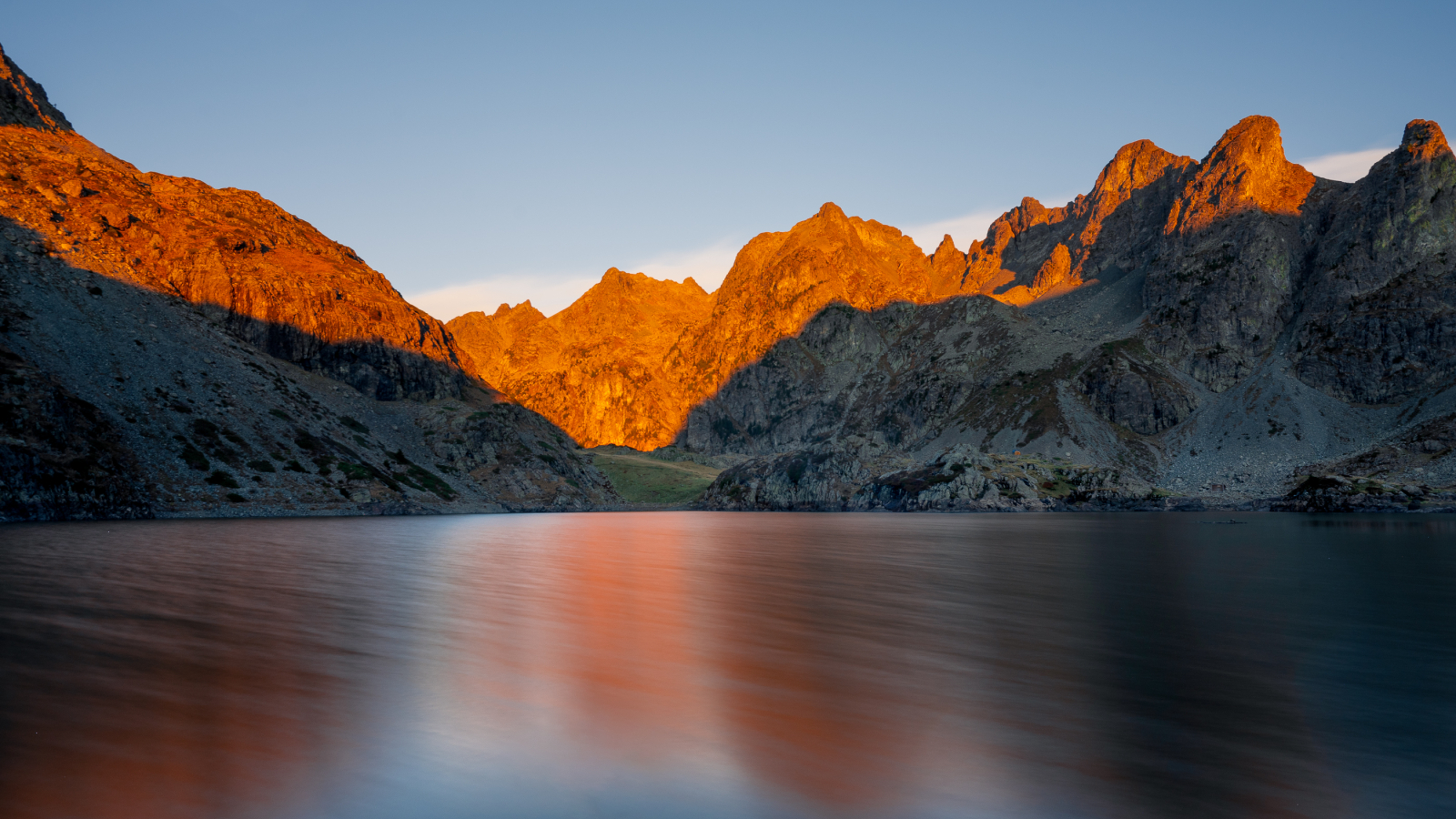 Un lac s'étend sous un ciel clair, avec des montagnes escarpées et irrégulières qui s'élèvent sur les côtés.