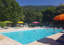 Une piscine rectangulaire est entourée d'une terrasse ensoleillée. Des parasols colorés sont plantés autour de la piscine, offrant de l'ombre aux nageurs. Des chaises longues sont disposées sur la terrasse. On distingue une pataugeoire ronde en arrière-plan.