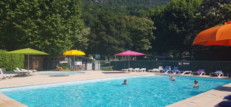 Une piscine rectangulaire est entourée d'une terrasse ensoleillée. Des parasols colorés sont plantés autour de la piscine, offrant de l'ombre aux nageurs. Des chaises longues sont disposées sur la terrasse. On distingue une pataugeoire ronde en arrière-plan.