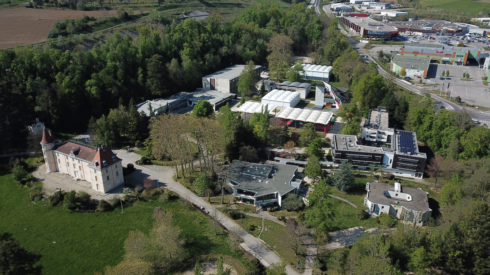 Une vue aérienne présente un grand campus aux multiples complexes, entouré d'arbres et bordé par une grande route reliée à un centre commercial