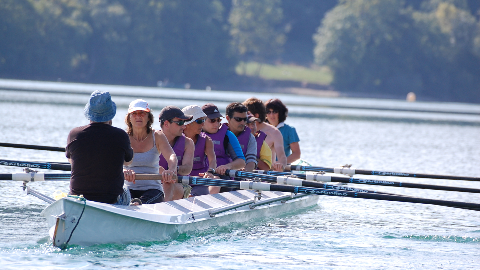 Sur les eaux cristallines du lac, une équipe de huit personnes rame sur un grand aviron, guidés par une personne qui leur donne le rythme. Au second plan, des arbres bordent les rives du lac.